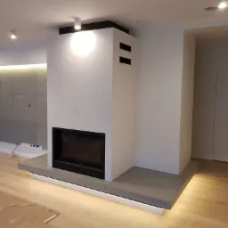 Nowoczesny salon z białym kominkiem obudowanym betonem architektonicznym, podświetlonym od dołu, z drewnianą podłogą zabezpieczoną kartonem podczas prac wykończeniowych.