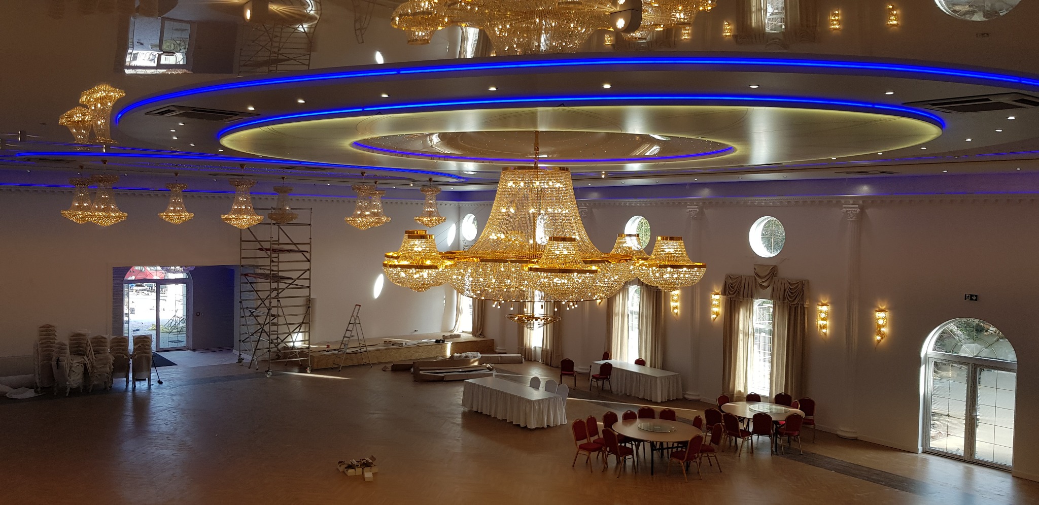 Przestronna sala balowa w trakcie remontu: widoczne rusztowania, kryształowe żyrandole, dekoracyjne oświetlenie sufitowe LED, przygotowane stoły i krzesła.