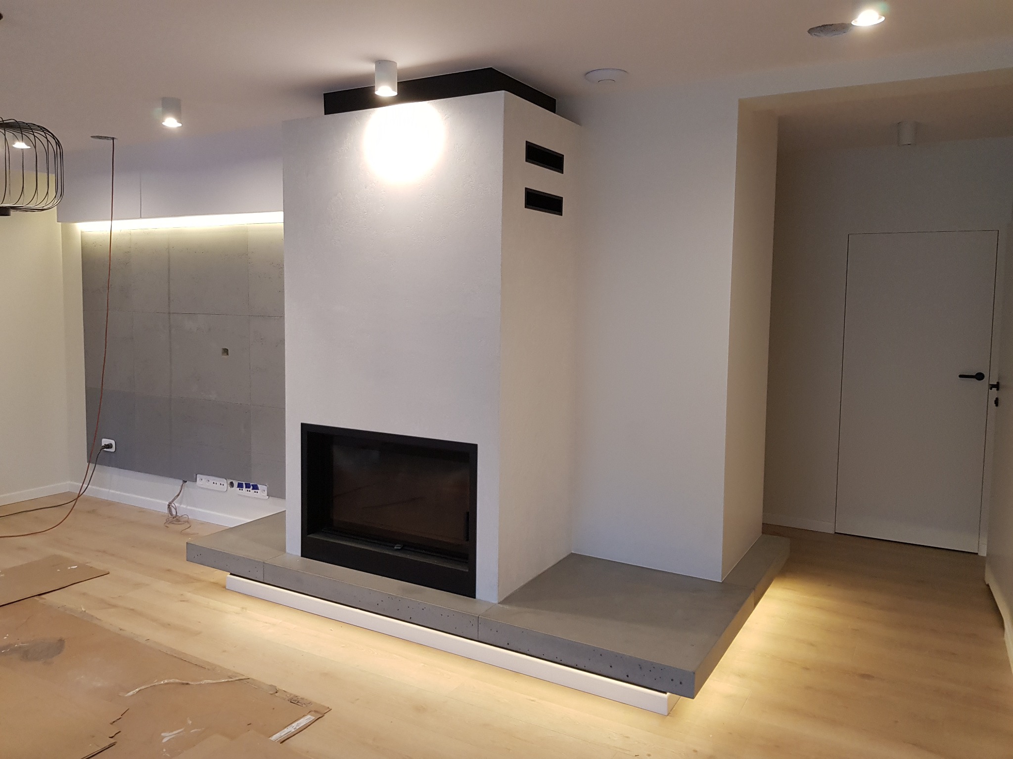 Nowoczesny salon z białym kominkiem obudowanym betonem architektonicznym, podświetlonym od dołu, z drewnianą podłogą zabezpieczoną kartonem podczas prac wykończeniowych.
