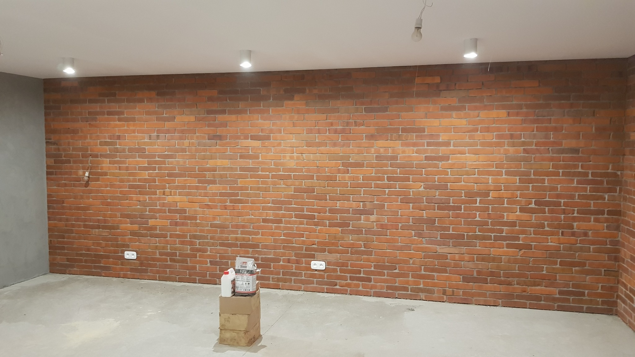 Wnętrze z wykończoną ścianą z cegły klinkierowej, widoczne puszki z fugą i gruntem na kartonach, surowy beton na podłodze i niedokończone oświetlenie sufitowe.