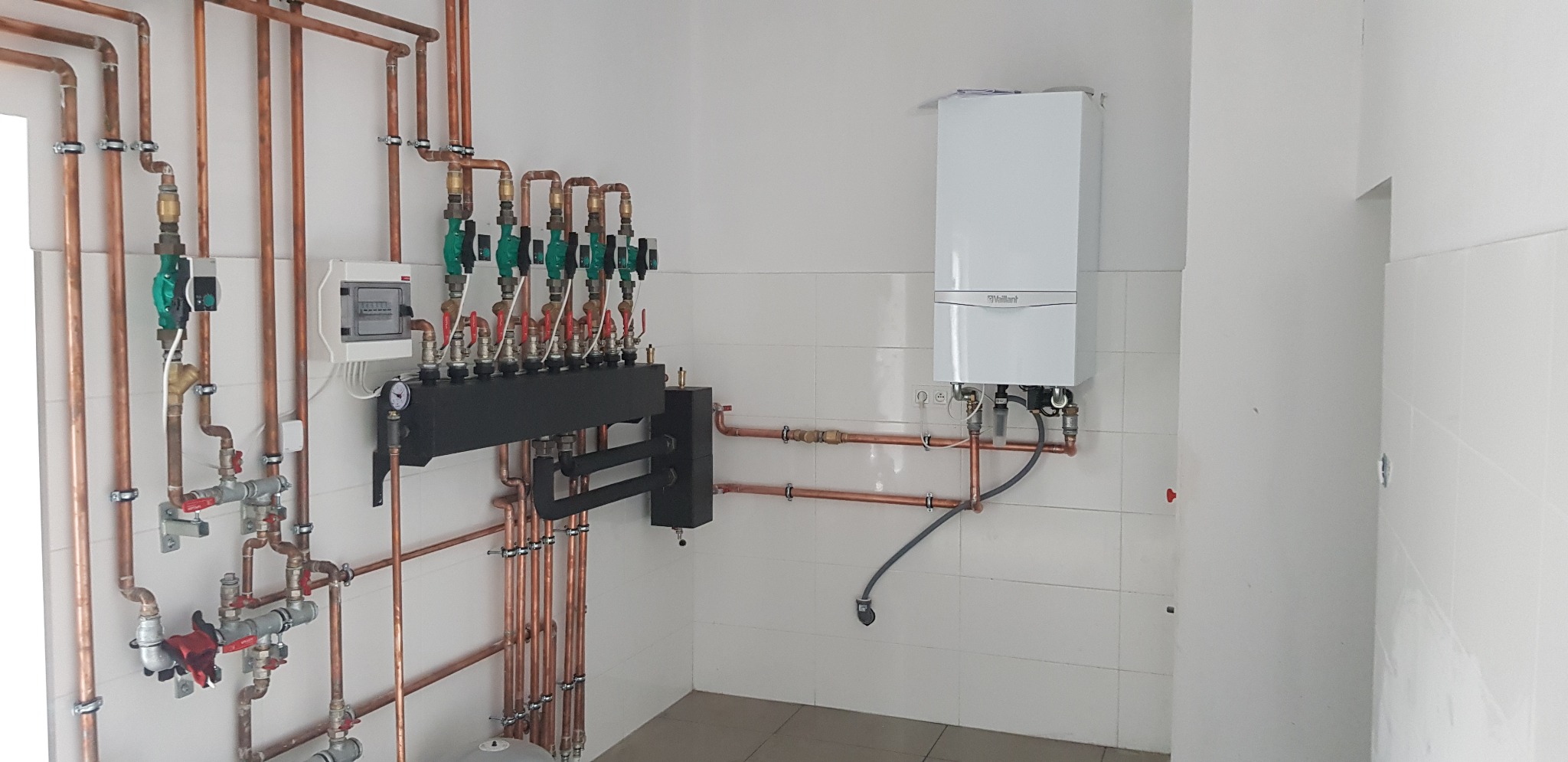 Nowoczesny, kondensacyjny piec gazowy Vaillant, podłączony do miedzianej instalacji centralnego ogrzewania z rozdzielaczem i licznikiem, zamontowany na ścianie z białych płytek.