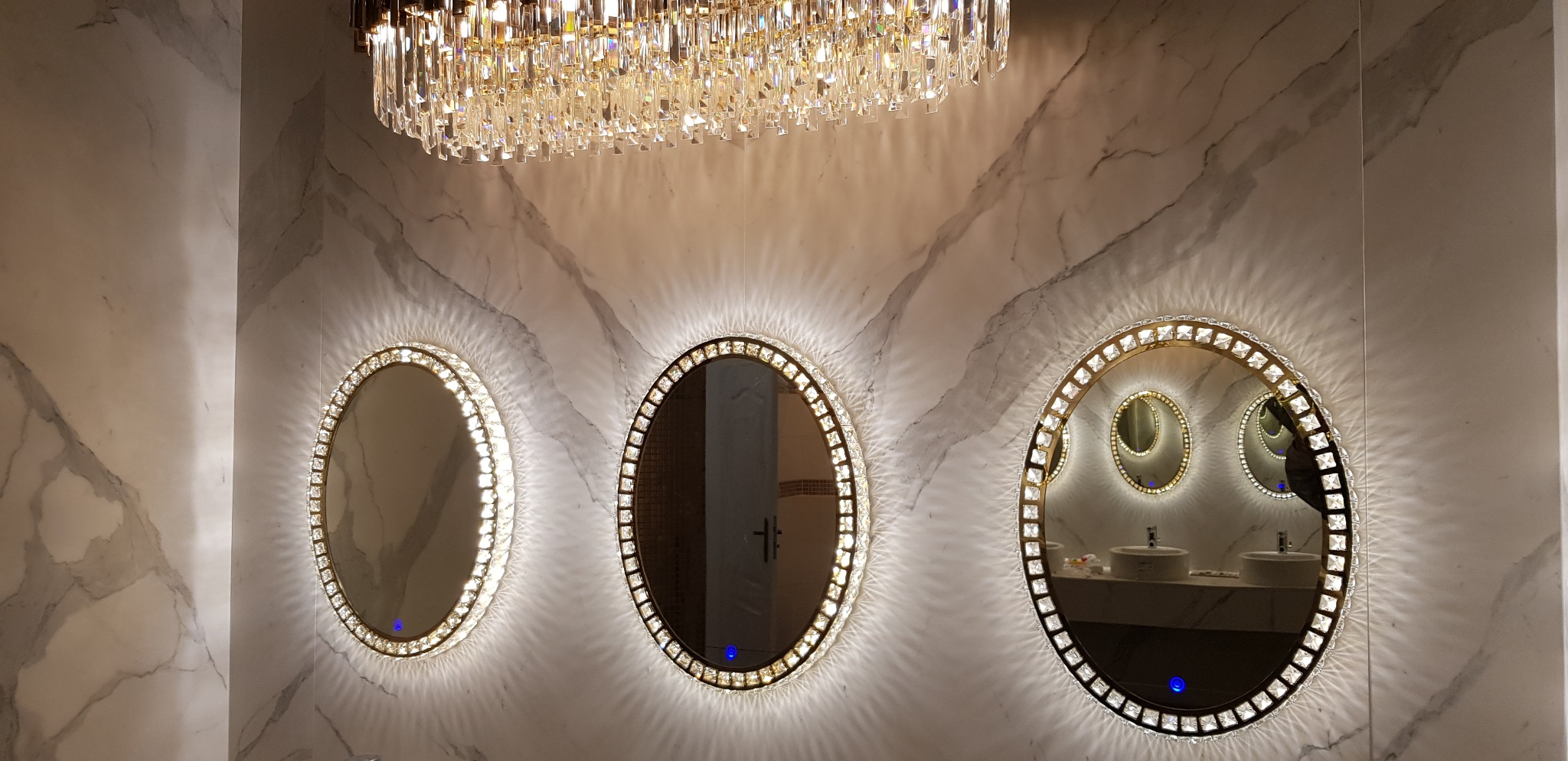 Nowoczesna łazienka z trzema owalnymi lustrami z podświetleniem LED i kryształowym żyrandolem na tle marmurowej ściany.