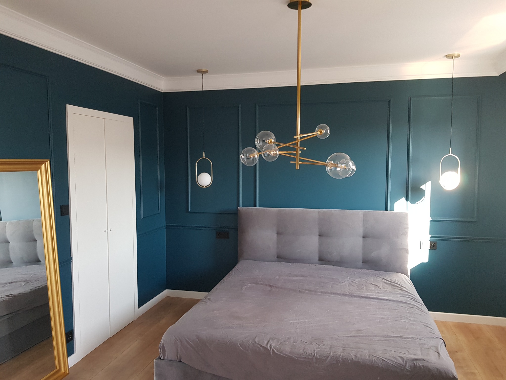 Sypialnia z granatowymi ścianami zdobionymi sztukaterią, szarym łóżkiem i złotymi lampami wiszącymi, widoczna biała szafa i lustro w złotej ramie.