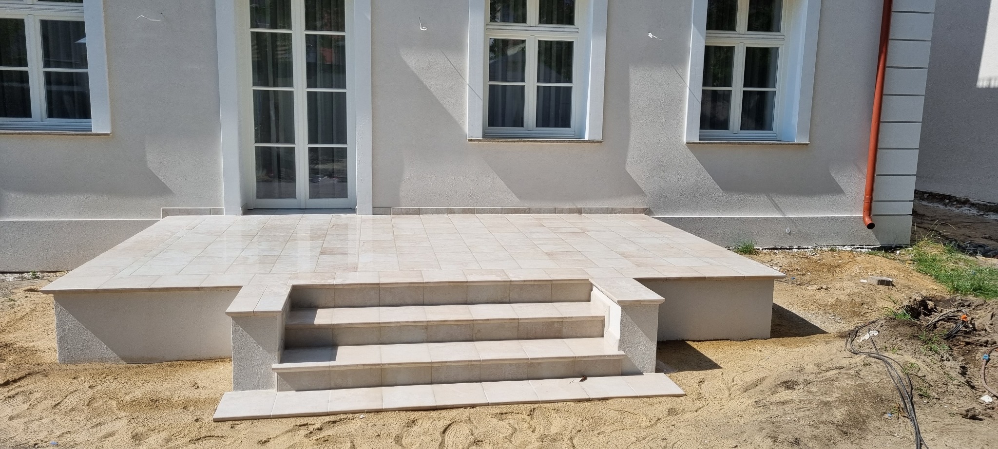 Wykończony taras z beżowych płytek z trzema schodami, dopasowany kolorystycznie do elewacji budynku z białymi oknami i drzwiami, widoczny pomarańczowy spust wody deszczowej.