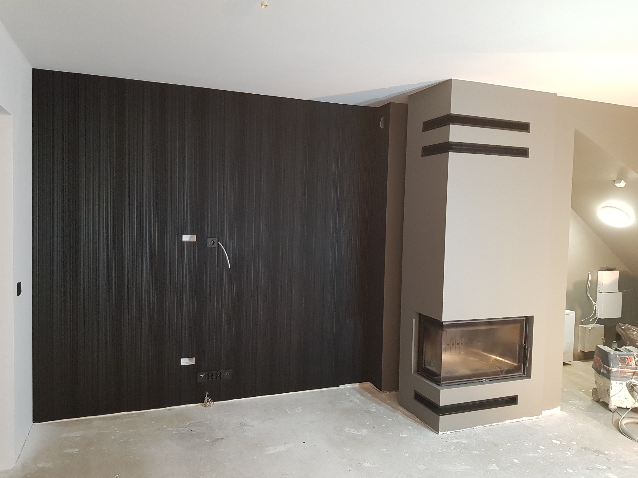 Pokój w trakcie wykańczania: ciemna ściana z pionowymi paskami, gniazdka elektryczne, szary kominek narożny z wkładem, surowa podłoga i sprzęt budowlany w tle.