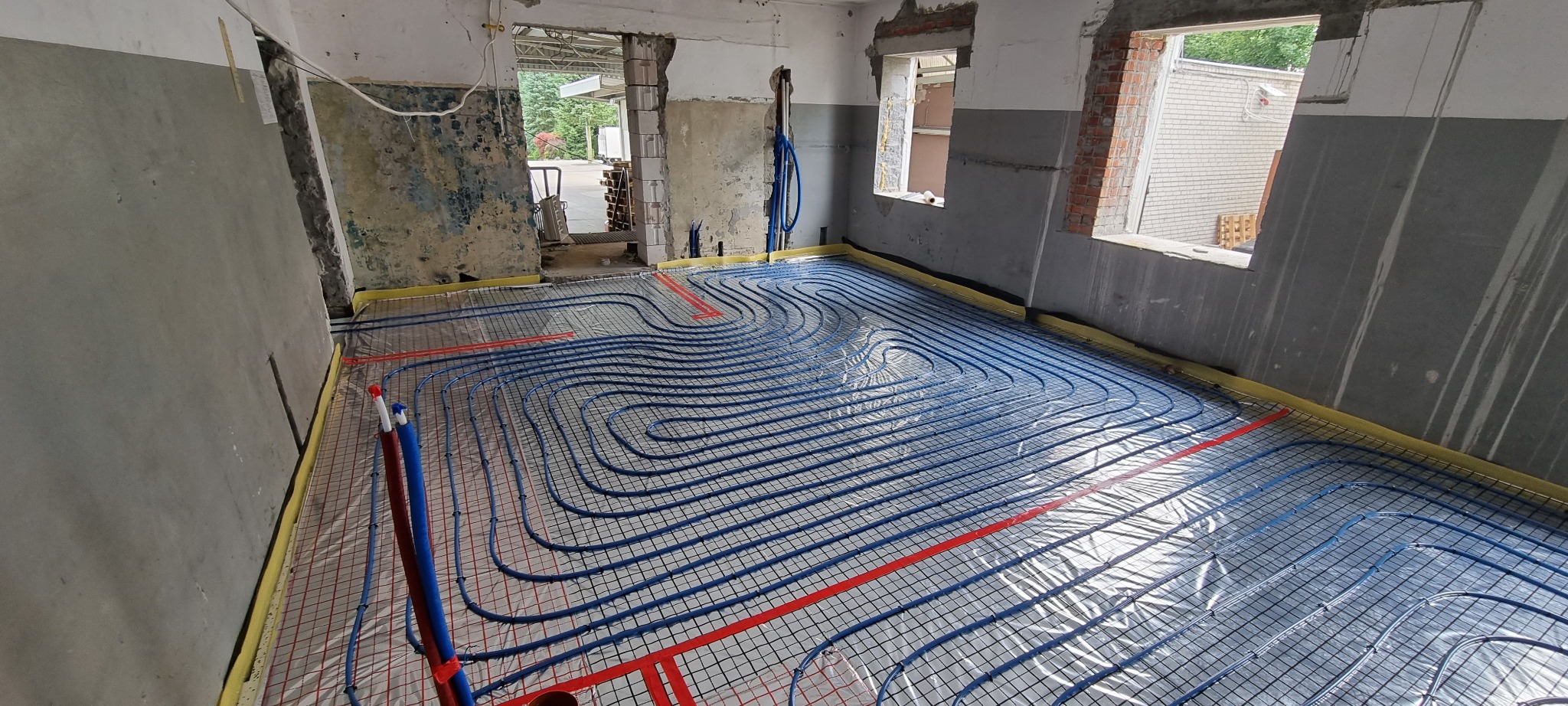 Niebieskie rurki ogrzewania podłogowego ułożone na siatce w surowym wnętrzu remontowanego pomieszczenia, widoczne okna i fragmenty instalacji hydraulicznej.