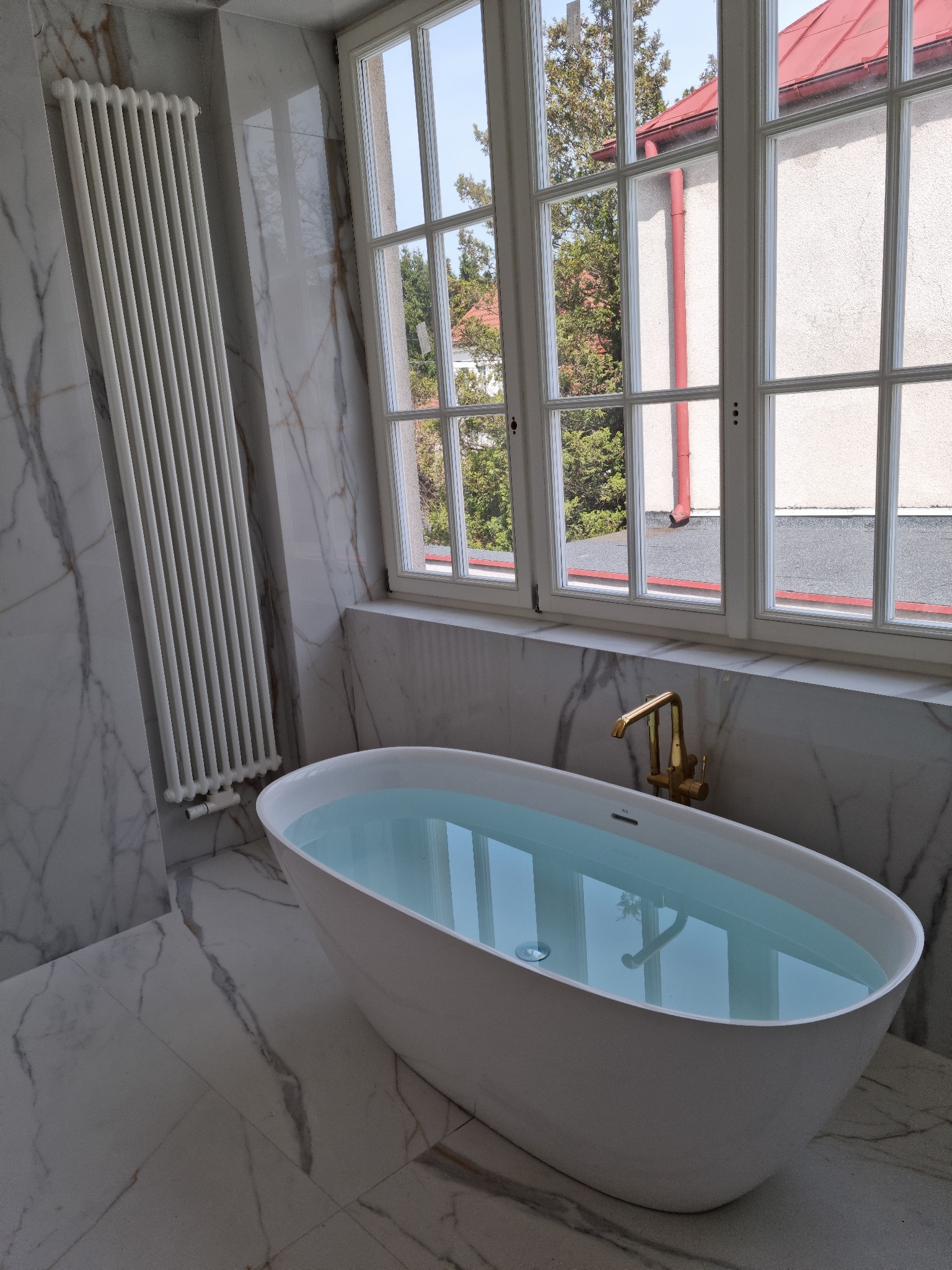 Elegancka, wolnostojąca wanna wypełniona wodą w łazience wykończonej marmurem, z widokiem na zewnątrz przez duże okno ze szprosami.