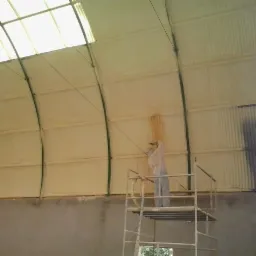 Pracownik w kombinezonie ochronnym na rusztowaniu natryskuje izolację na wewnętrzną stronę dachu hali, widoczne fragmenty już pokryte pianką oraz elementy konstrukcyjne dachu.