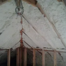 Poddasze ocieplone białą pianką, widoczne drewniane elementy konstrukcyjne, ujęcie z dołu w trakcie prac, szare przewody zwisające z sufitu.