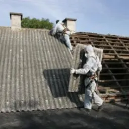 Dwóch pracowników w kombinezonach ochronnych zdejmuje płyty eternitu z dachu domu, widoczna konstrukcja drewniana dachu oraz komin.