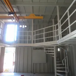 Wnętrze kontenera po adaptacji z antresolą, spiralnymi schodami i suwnicą pomalowane na biało.