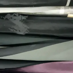 Stos pofałdowanych tkanin w odcieniach czerni, szarości i fioletu, z widocznymi fakturami i delikatnym połyskiem.