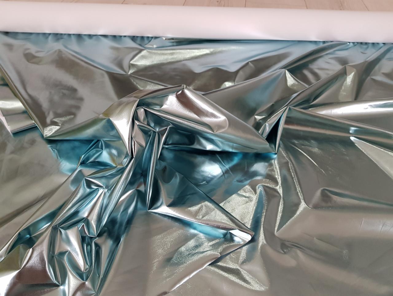 Błyszcząca, turkusowa tkanina metaliczna w fałdach, zwinięta na białym wałku.