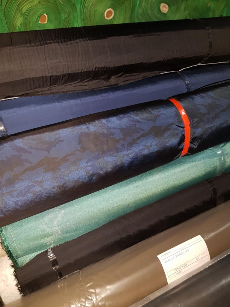 Stos zwiniętych bel materiałów o różnej kolorystyce i fakturze, widoczne etykiety na niektórych rolkach, ułożone pod kątem.