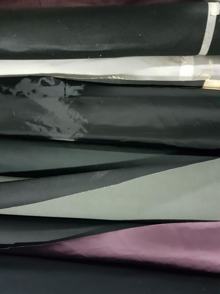 Stos pofałdowanych tkanin w odcieniach czerni, szarości i fioletu, z widocznymi fakturami i delikatnym połyskiem.