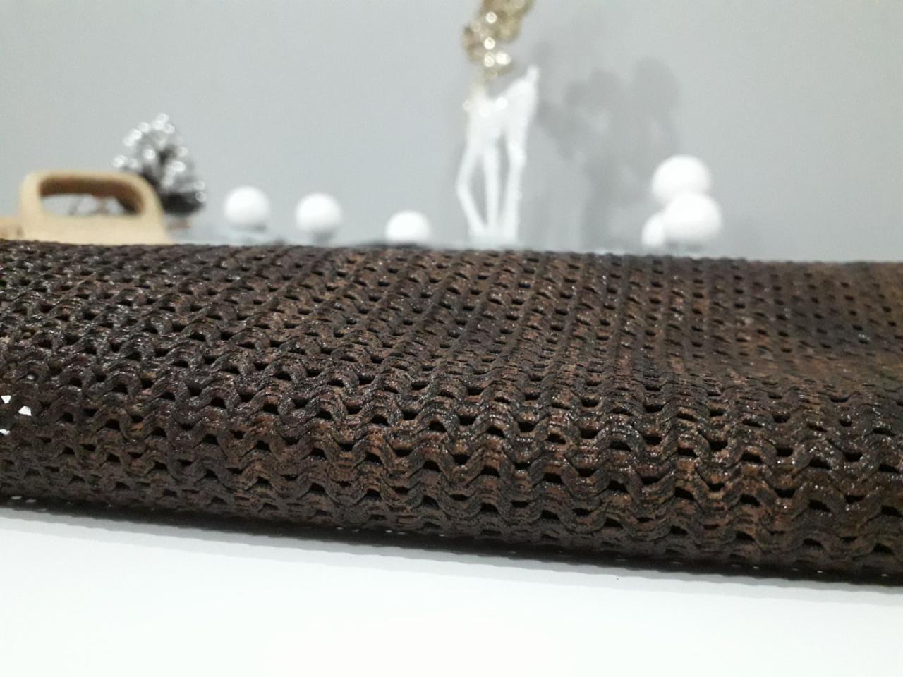 Ciemnobrązowa, ażurowa tkanina o wyraźnej strukturze, leżąca na białym blacie, z rozmytym tłem dekoracji świątecznych.