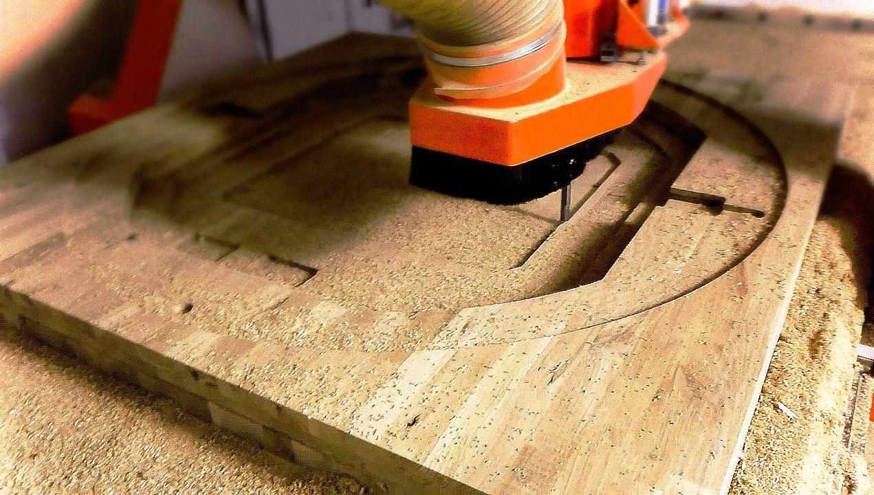 Frezarka CNC wycina okrągły kształt w drewnianej płycie, wióry i pył na powierzchni.