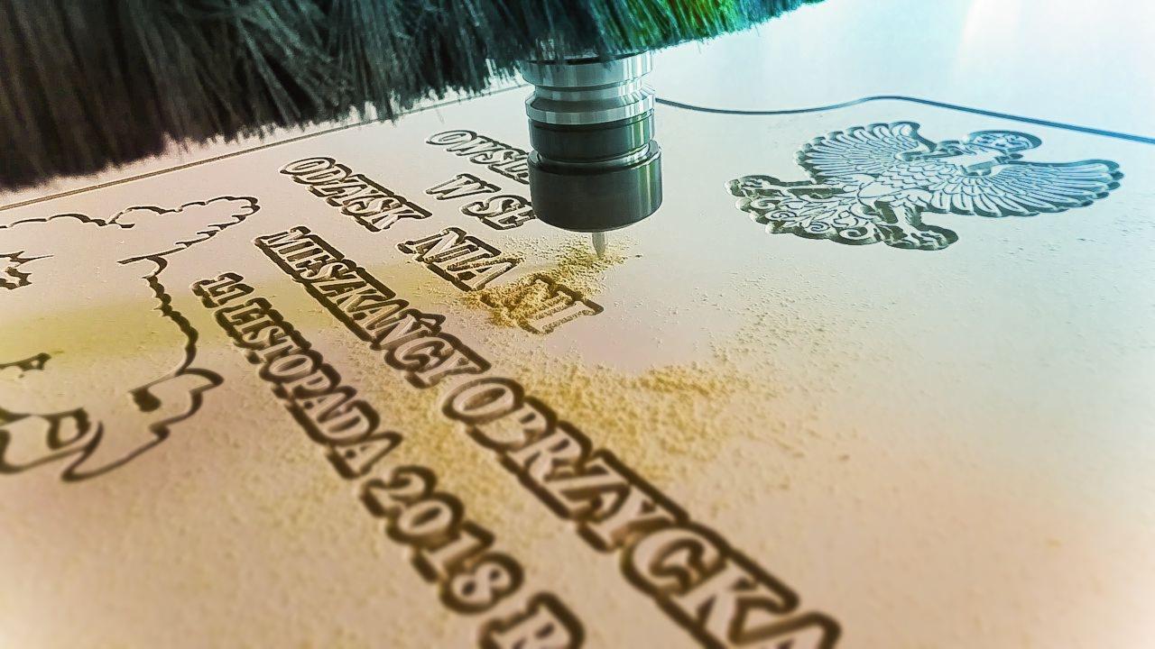 Frezarka CNC wycina w drewnie napis 'Odznak W Służbie Mieszkańcy Obrazyccy 11 Listopada 2018 R' oraz orła w koronie, widoczne drobne wióry.