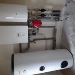 Kocioł grzewczy, zasobnik ciepłej wody i czerwony zbiornik wyrównawczy połączone rurami w kotłowni; widoczne przewody z izolacją termiczną. Ściana z fragmentem płyty Swiss Krono.