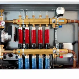 Rozdzielacz ogrzewania podłogowego w metalowej szafce, widoczne mosiężne rury, czerwone i niebieskie pętle grzewcze, manometr, pompa obiegowa Wilo i regulator temperatury, z logo producenta...