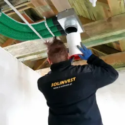 Pracownik w kurtce z logo firmy łączy biały kanał wentylacyjny z metalową skrzynką pod sufitem z drewnianych belek, widoczne zielone rury karbowane.