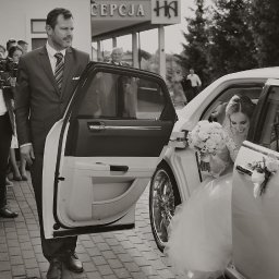 FOTO VISION WOJCIECH CHUDZIK - Czarno-białe zdjęcie panny młodej uśmiechającej się w limuzynie z otwartymi drzwiami, obok kamerzysta z gimbalem i mężczyzna w garniturze.