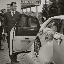 Czarno-białe zdjęcie panny młodej uśmiechającej się w limuzynie z otwartymi drzwiami, obok kamerzysta z gimbalem i mężczyzna w garniturze.