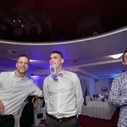 FOTO VISION WOJCIECH CHUDZIK - Trzech uśmiechniętych mężczyzn w eleganckich strojach w sali bankietowej. Jeden z nich trzyma muszkę w ustach.