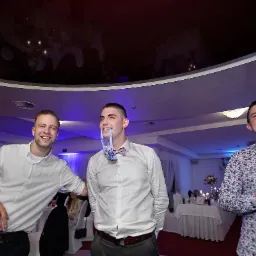 Trzech uśmiechniętych mężczyzn w eleganckich strojach w sali bankietowej. Jeden z nich trzyma muszkę w ustach.