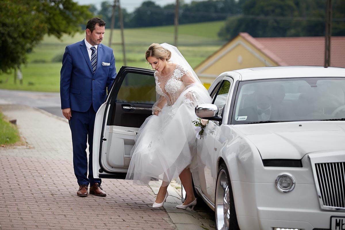 Panna młoda w koronkowej sukni wysiada z białej limuzyny, obok stoi mężczyzna w granatowym garniturze.