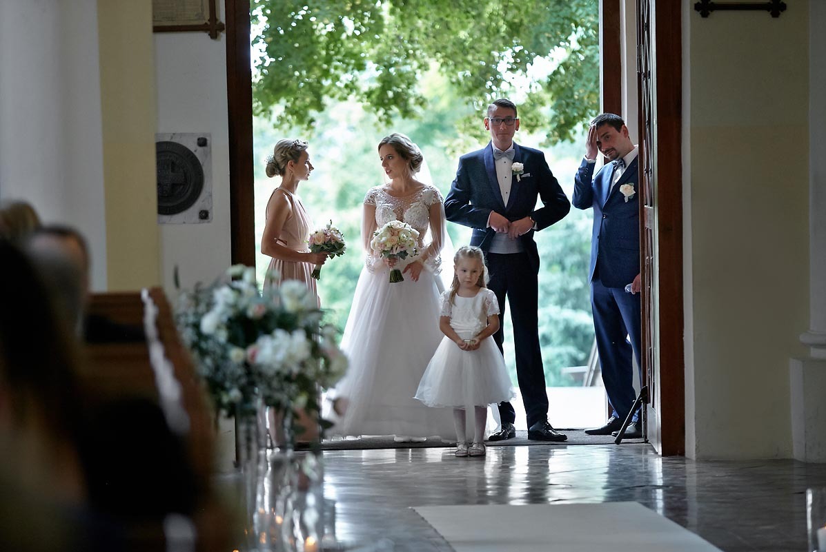 Panna młoda w koronkowej sukni, pan młody w smokingu, druhna z bukietem i dziewczynka w białej sukience stoją w wejściu do kościoła, widok z wnętrza kościoła, w tle zieleń.