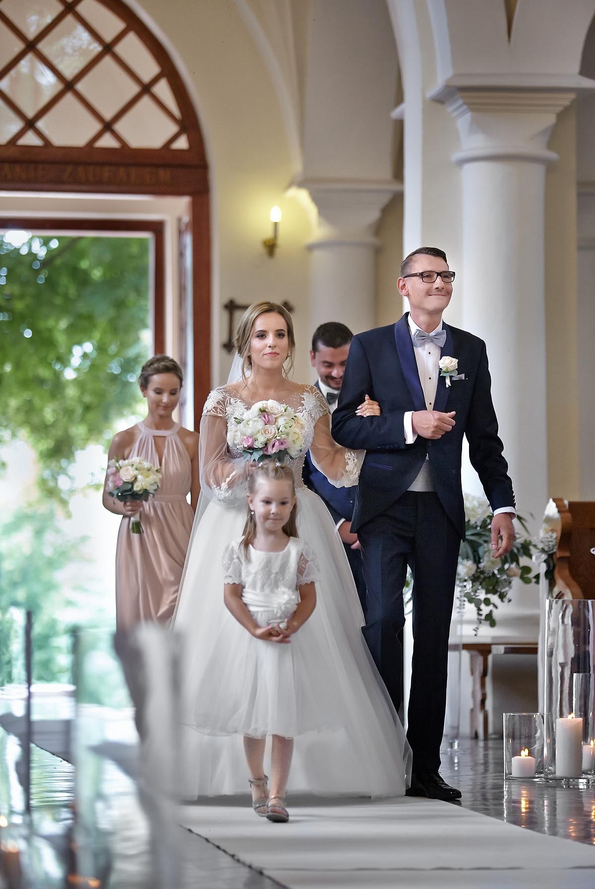 Uśmiechnięty pan młody w granatowym smokingu prowadzi pannę młodą w białej sukni i welonie do ołtarza, za nimi druhna i mała dziewczynka w białej sukience, w tle kolumny i świece.