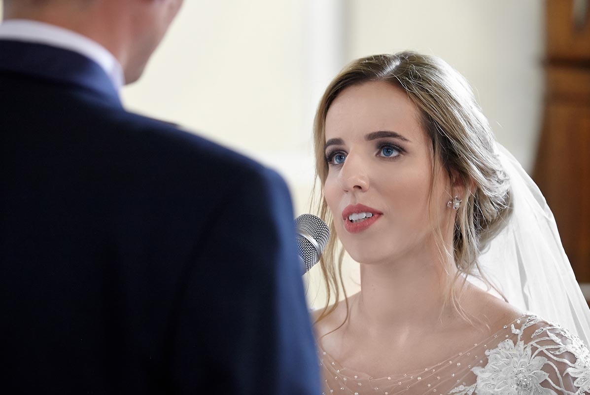 Panna młoda w sukni ślubnej z koronkowymi zdobieniami, trzymająca mikrofon i składająca przysięgę małżeńską, z widocznym fragmentem garnituru pana młodego.