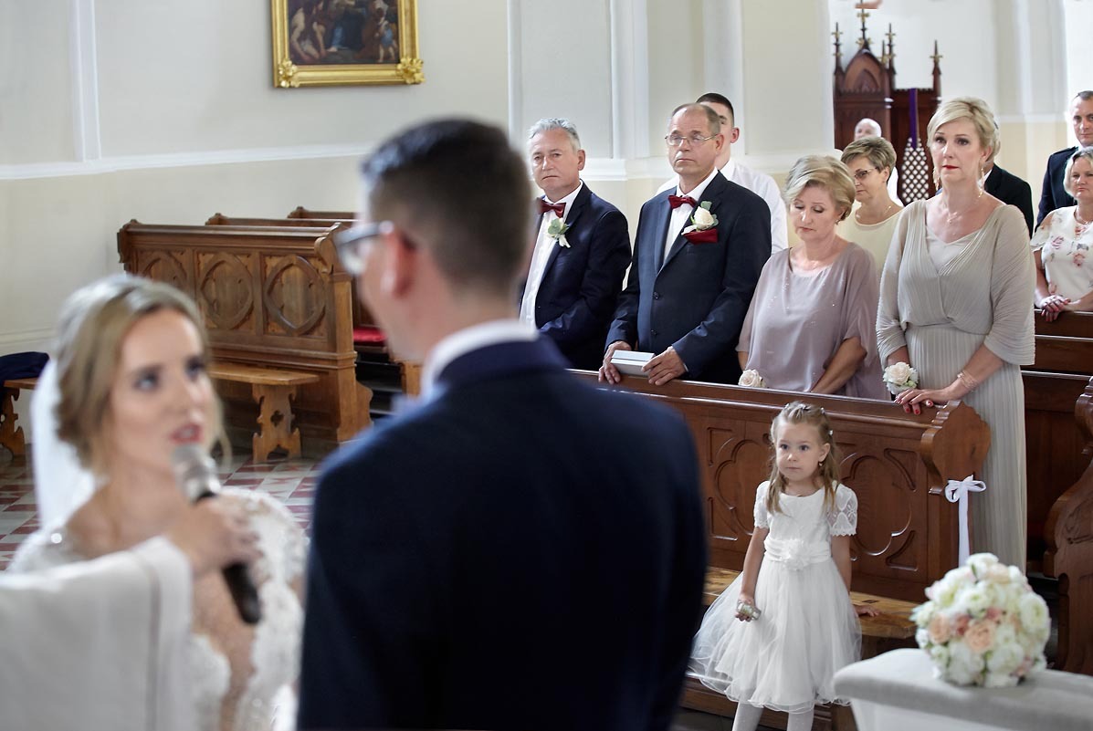 Panna młoda z mikrofonem wygłasza przysięgę małżeńską w kościele, w tle goście weselni i dziewczynka w białej sukience.