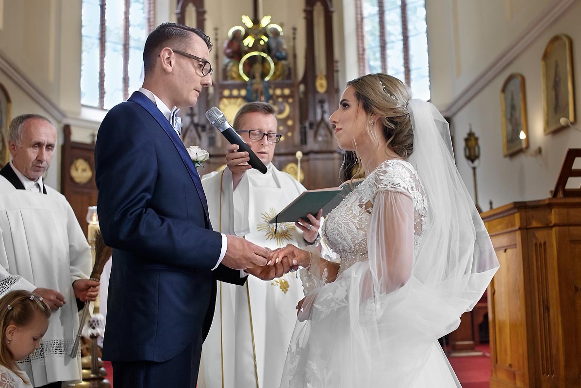 Para młoda podczas ceremonii zaślubin w kościele, wymiana obrączek, ksiądz w tle, panna młoda w koronkowej sukni z welonem, pan młody w granatowym garniturze.