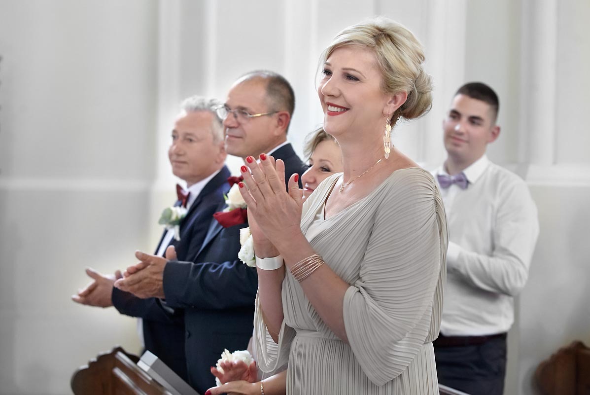 Goście weselni klaszczą podczas ceremonii, panna młoda w beżowej sukni z plisowanymi rękawami i złotymi kolczykami.