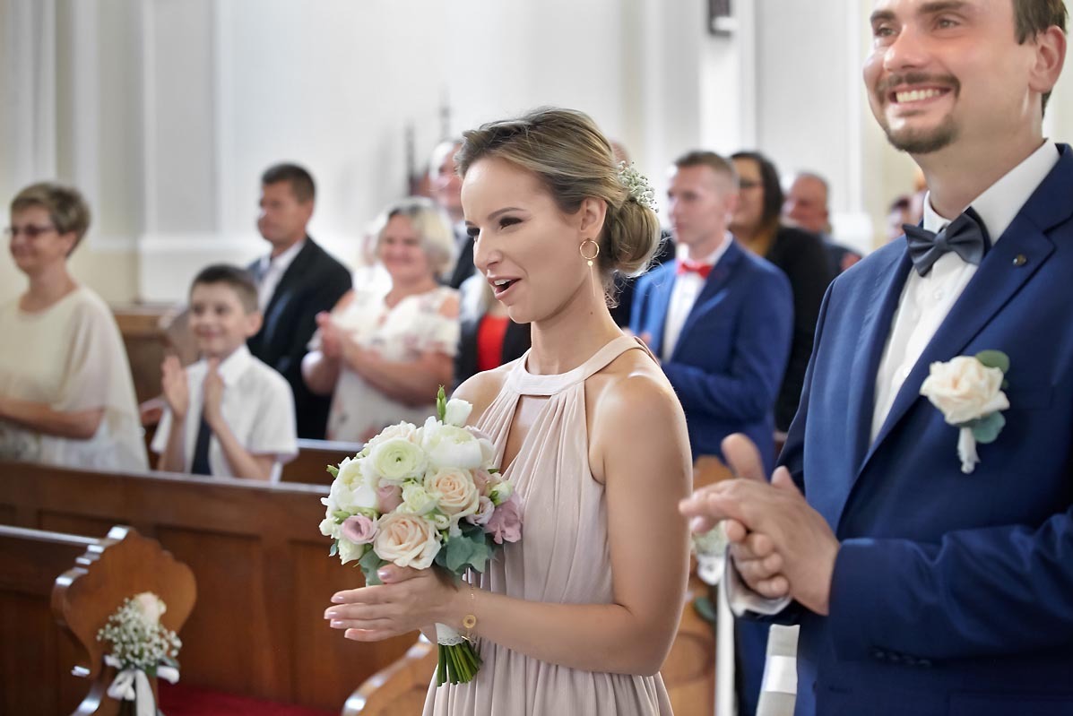 Uśmiechnięta druhna w sukience w kolorze pudrowego różu trzyma bukiet białych i różowych kwiatów podczas ceremonii ślubnej w kościele, w tle goście biją brawo.