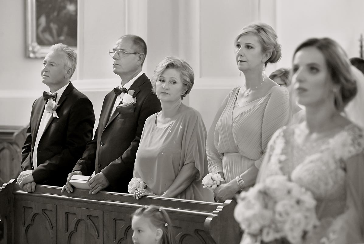 Czarno-białe zdjęcie gości weselnych w kościele, w pierwszym rzędzie Panna Młoda z bukietem, za nią rodzina, w tle obraz na ścianie.