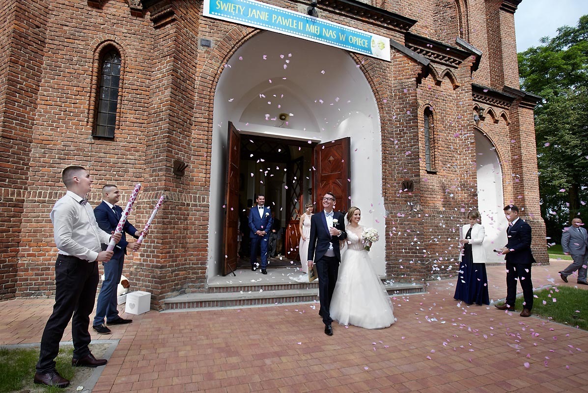 Para młoda wychodzi z kościoła obsypana płatkami róż, w tle goście i mężczyźni z tubami konfetti.