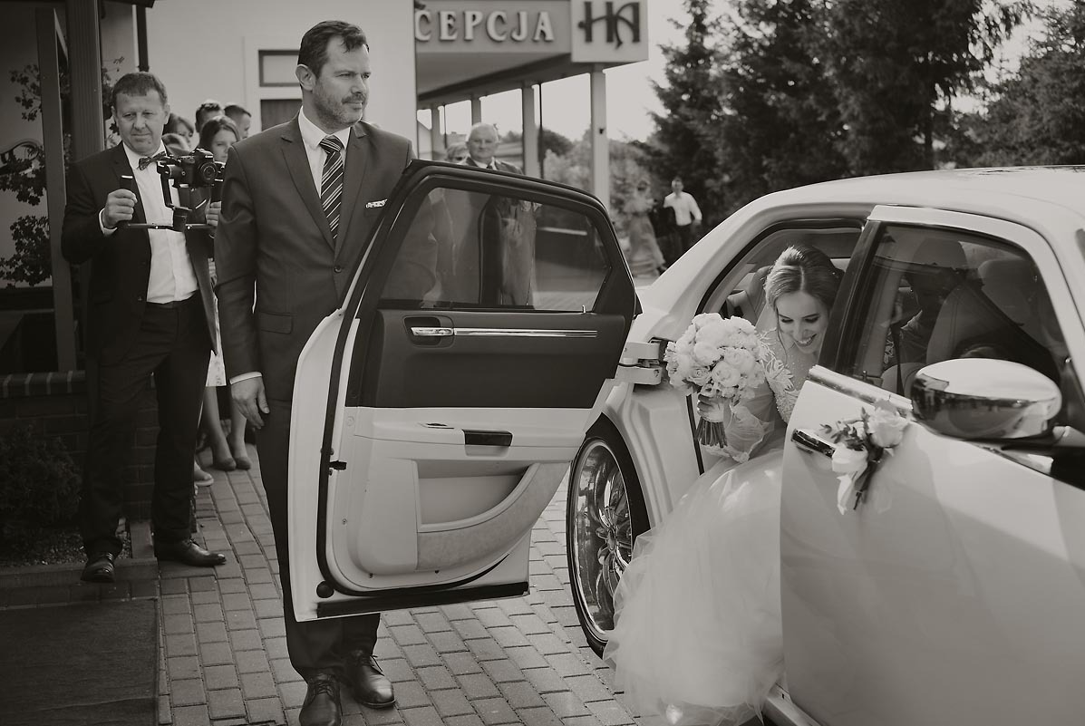 Czarno-białe zdjęcie panny młodej uśmiechającej się w limuzynie z otwartymi drzwiami, obok kamerzysta z gimbalem i mężczyzna w garniturze.