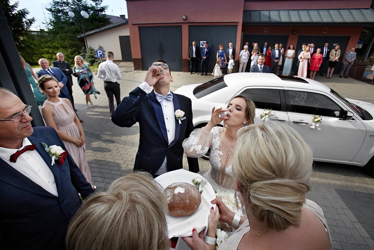 Para młoda w trakcie wznoszenia toastu wódką przed białym samochodem udekorowanym kwiatami, w tle goście weselni i budynek z garażami.