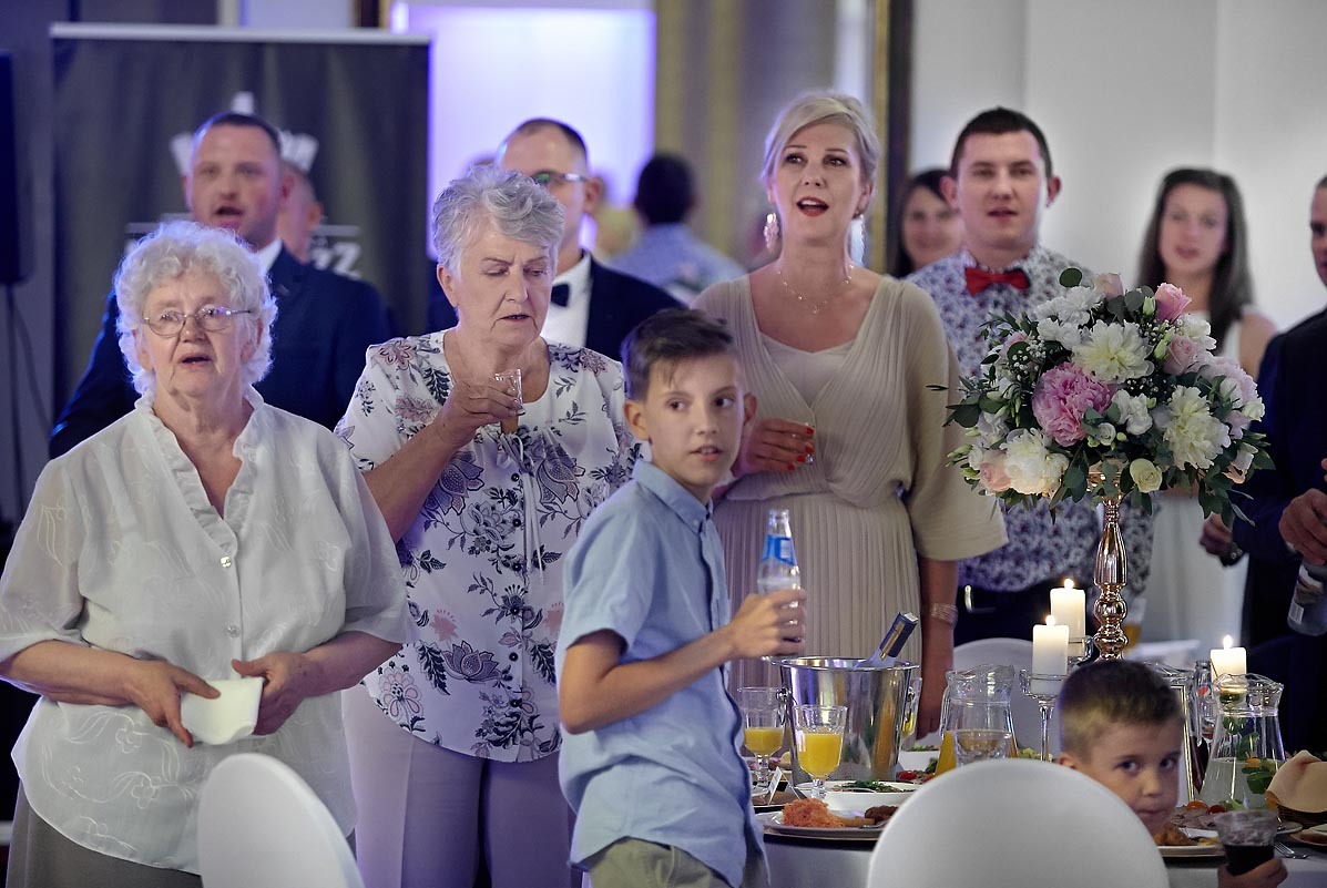 Goście weselni w trakcie toastu, w tle dekoracje kwiatowe i świece na stole.