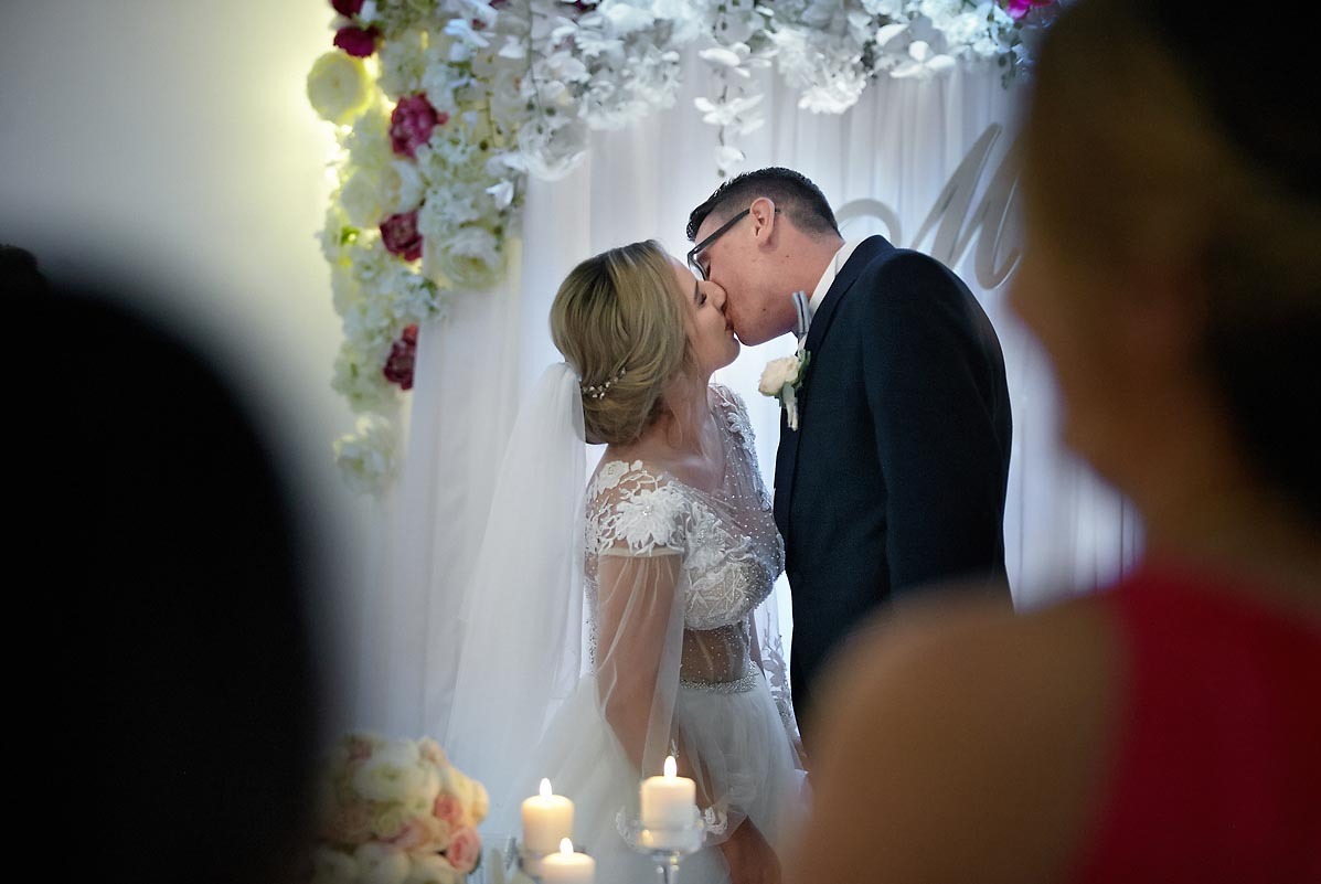 Pocałunek nowożeńców pod kwiatową dekoracją podczas ceremonii ślubnej, widoczne świece i fragmenty gości w tle.