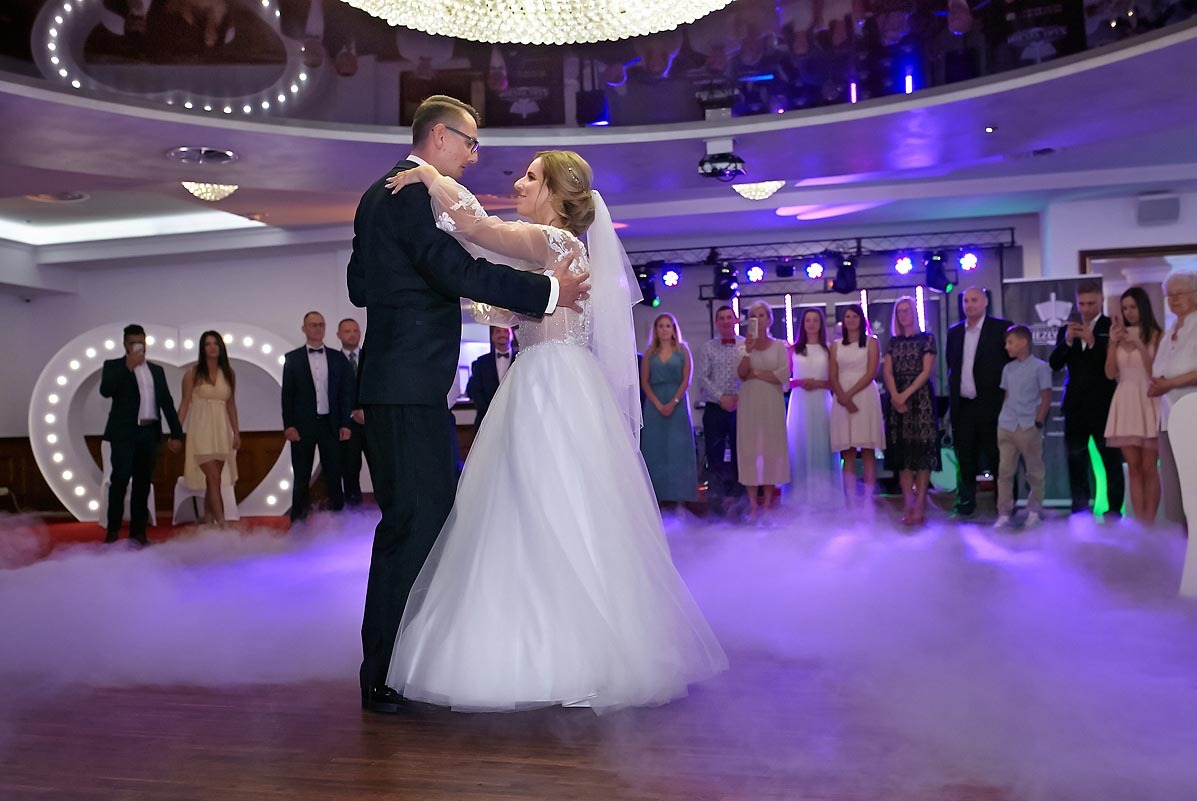 Para młoda tańczy pierwszy taniec na tle dekoracji w kształcie serca i efektu dymu, goście weselni obserwują.