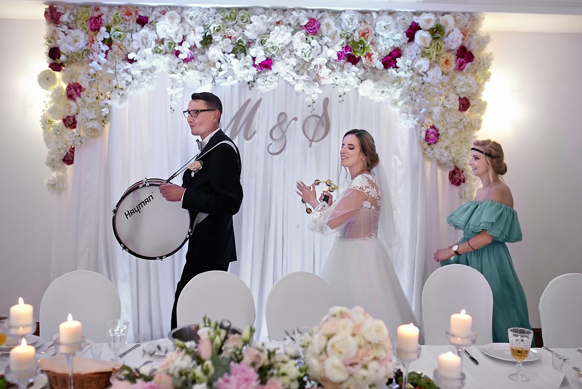 Para młoda świętuje wesele z bębniarzem i druhną na tle kwiatowej dekoracji i stołu z płonącymi świecami.