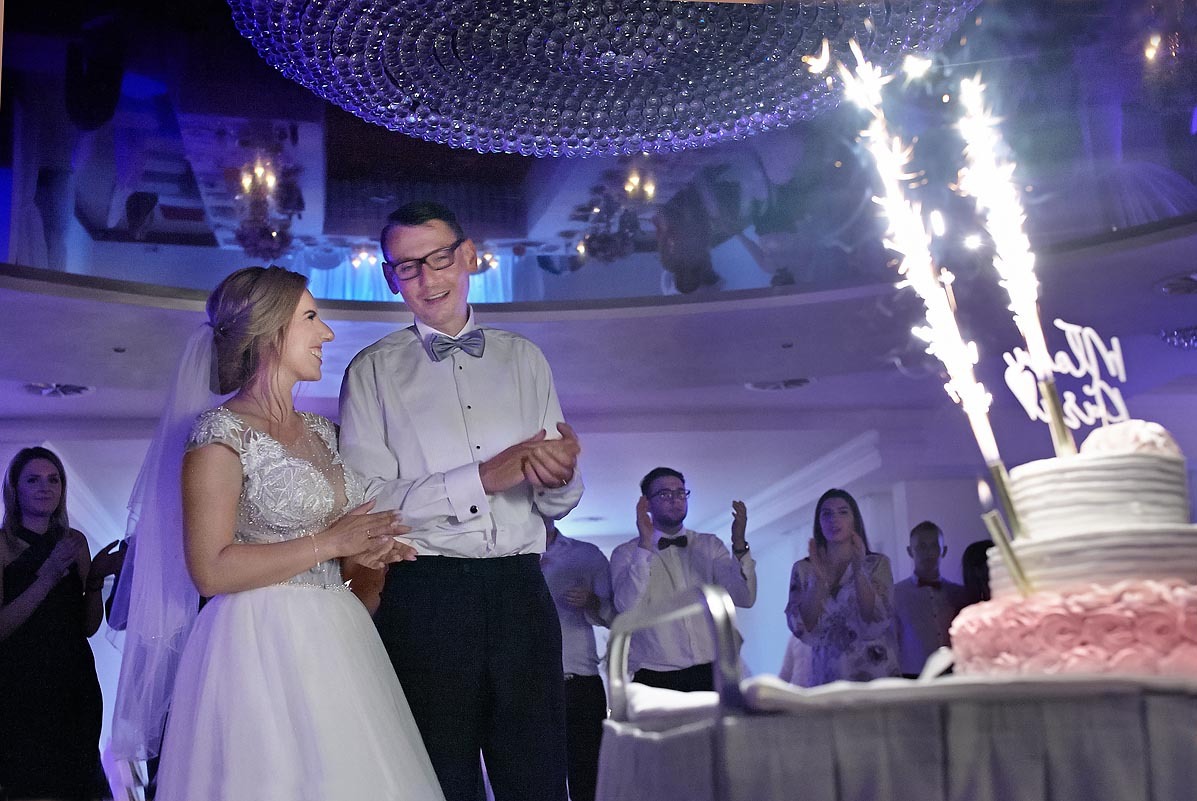 Uśmiechnięta para młoda w trakcie krojenia tortu weselnego z fontannami iskier, goście w tle klaszczą.