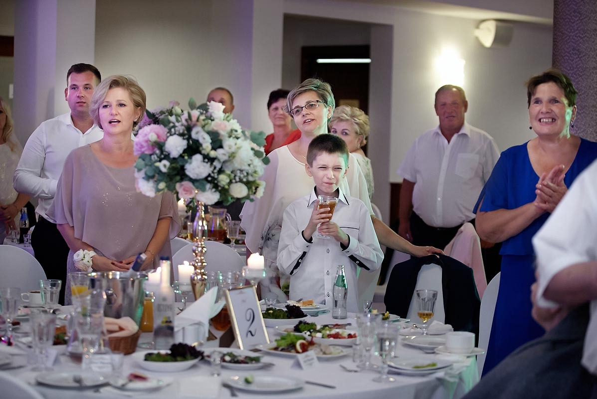 Uroczysty stół z dekoracją kwiatową, świecami i zastawą, w tle goście weselni, chłopiec pije sok z uśmiechem.