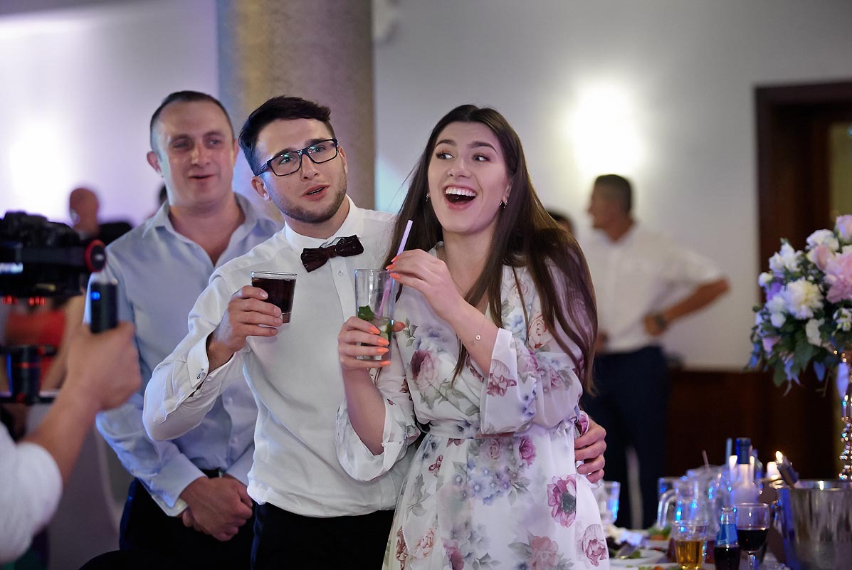 Uśmiechnięta para młoda z gośćmi pozuje z drinkami podczas wesela, w tle widoczny kamerzysta z profesjonalnym sprzętem.