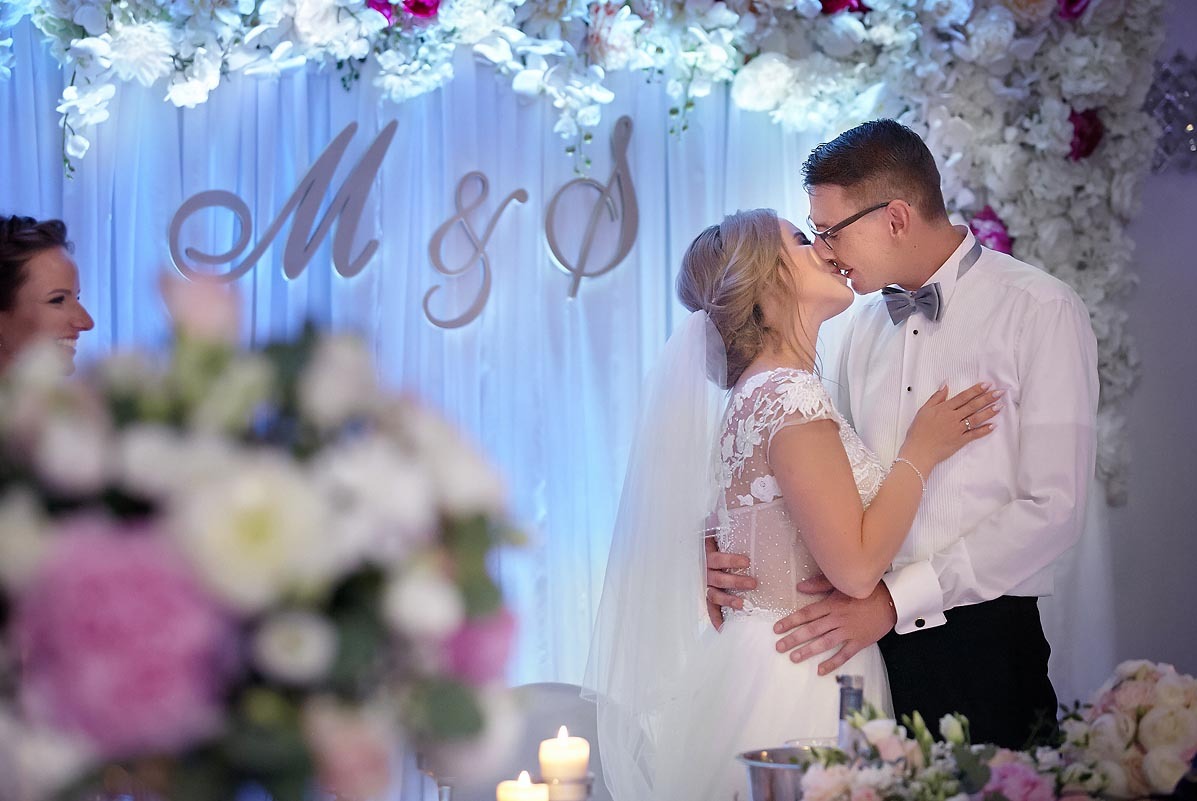 Pocałunek młodej pary na tle dekoracji ślubnej z białych i różowych kwiatów oraz podświetlanych liter 'M & S', z widocznym bukietem na pierwszym planie i uśmiechniętą kobietą w tle.