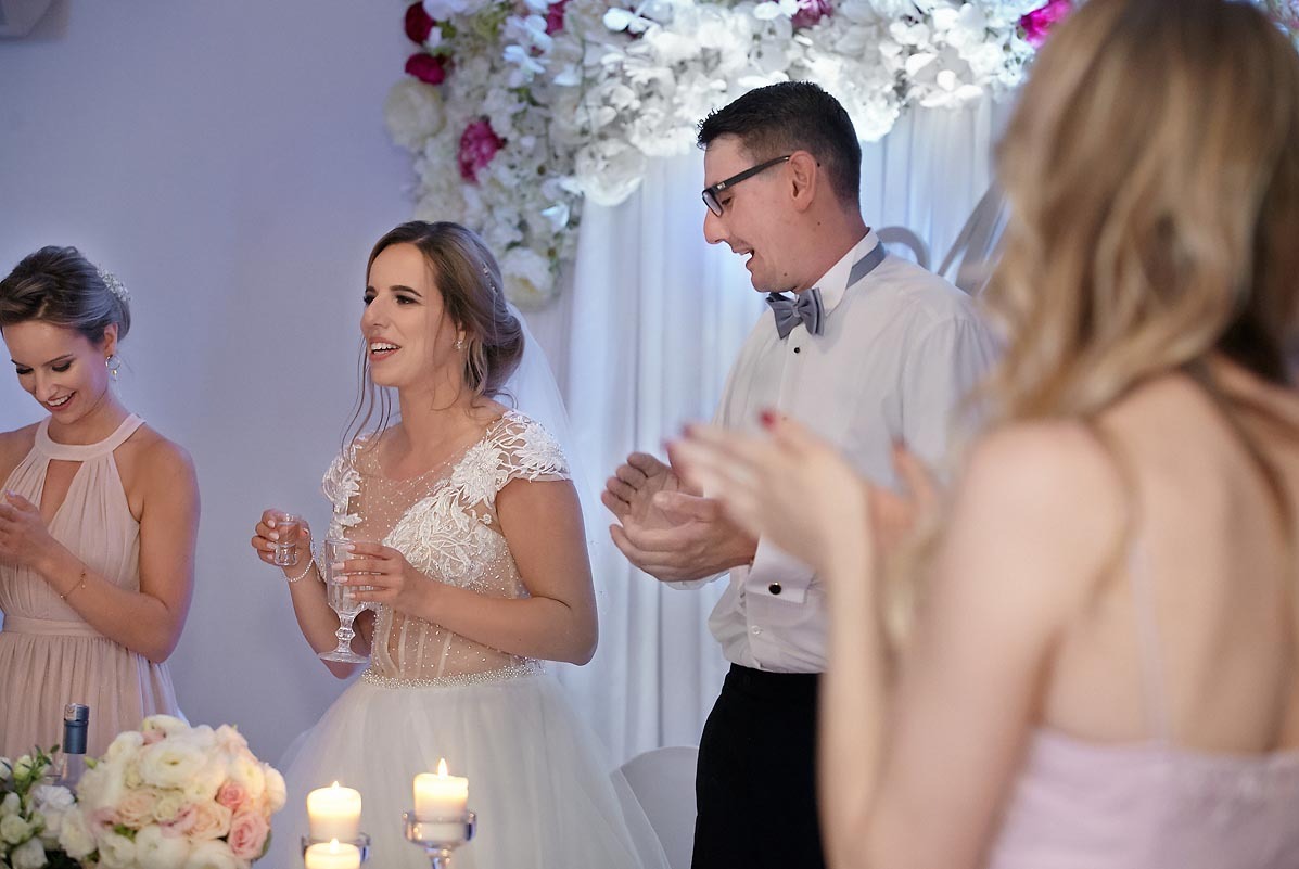 Panna młoda w koronkowej sukni, toast weselny z gośćmi, dekoracja kwiatowa w tle.